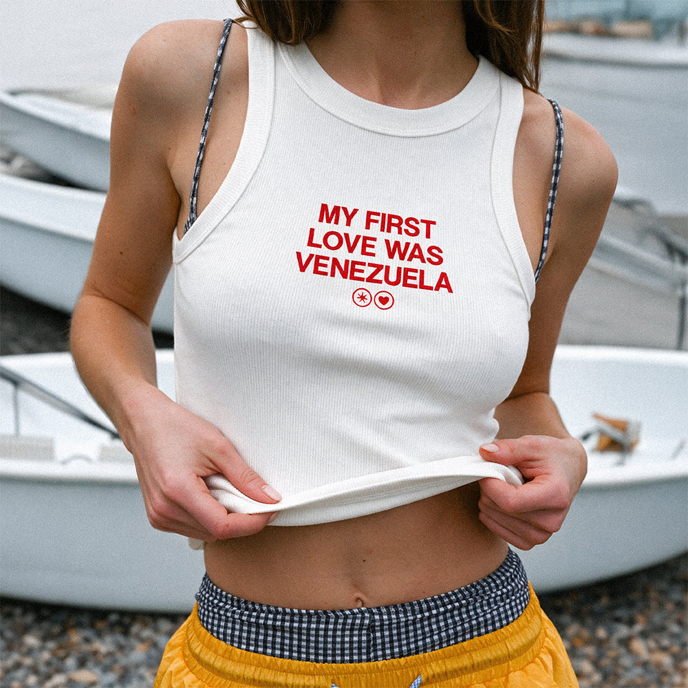 Mujer llevando la camiseta de tirantes blanca «My First Love Was Venezuela» de Caracas Merch, con barcas al fondo